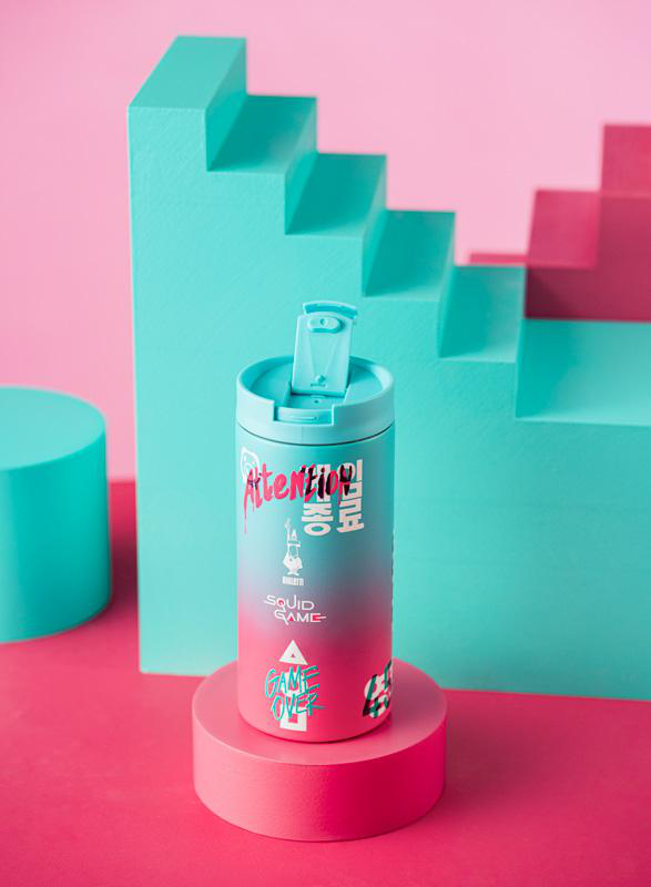 Ein Squid Game-Tumbler auf einem rosa und blaugrünen Hintergrund.