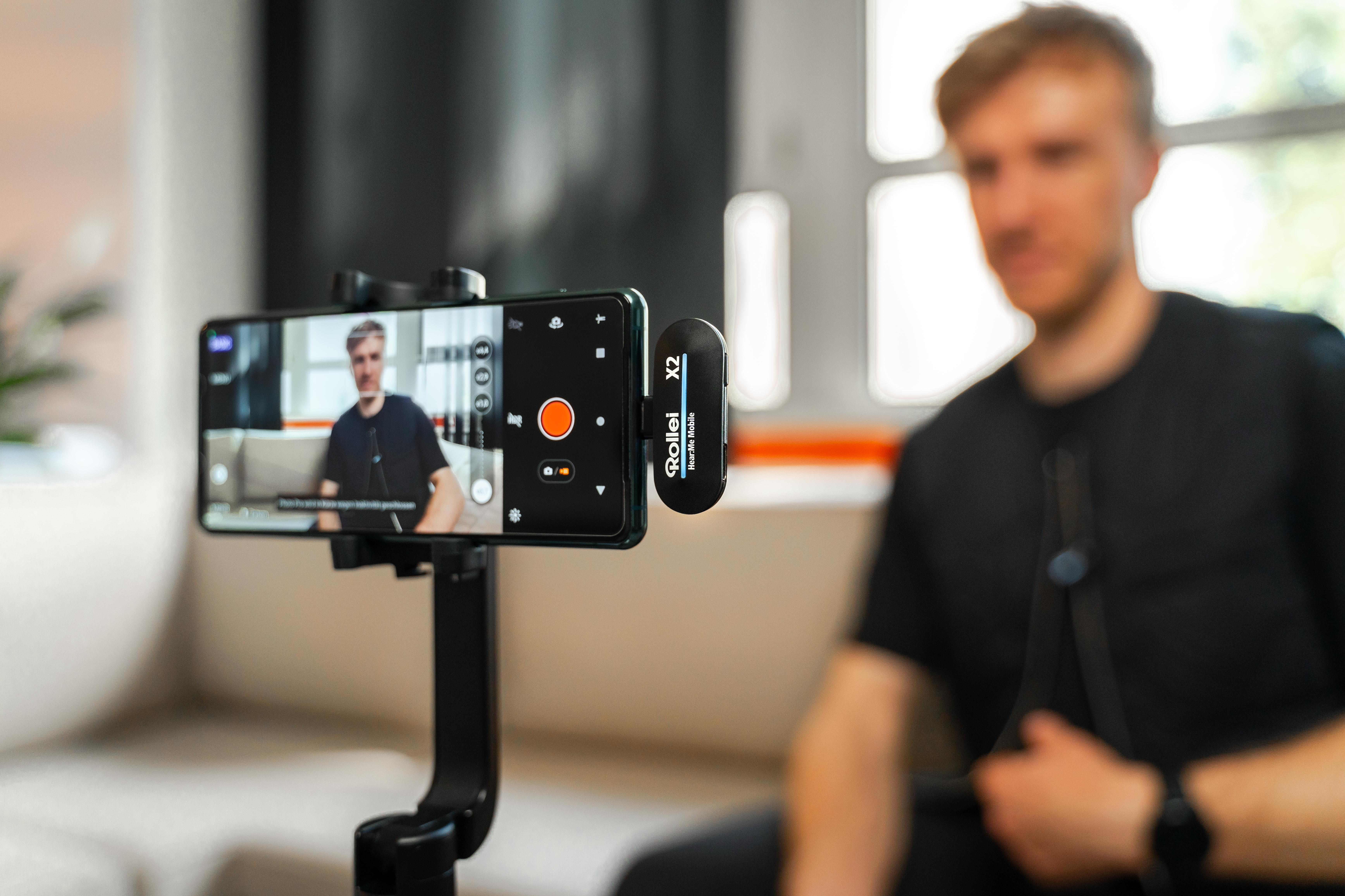 Ein Mann filmt sich selbst mit einem auf einem Stativ befestigten Smartphone. Ein kleines Mikrofon ist an seinem Hemd befestigt.