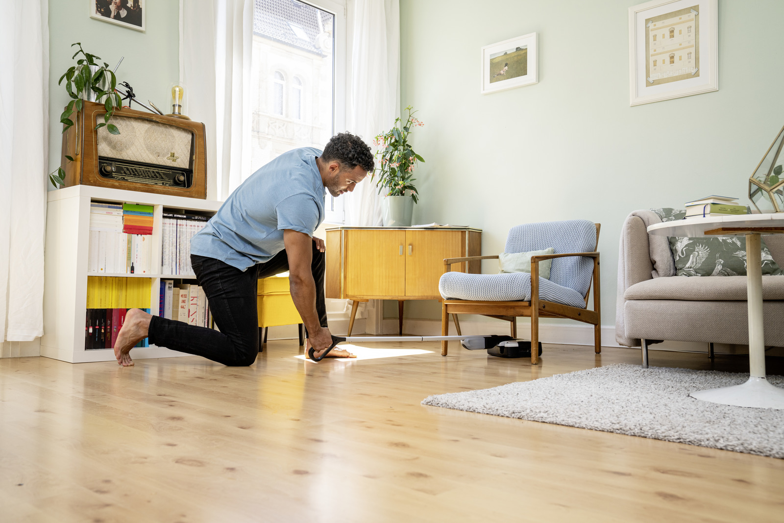 Mężczyzna klęczy, czyszcząc podłogę odkurzaczem robota. W tle pokój z meblami.