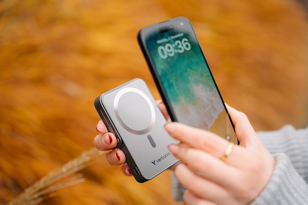 Kéz egy telefont tart egy ezüst Verbatim power bank mellett. A telefonon az idő 09:36 látható.