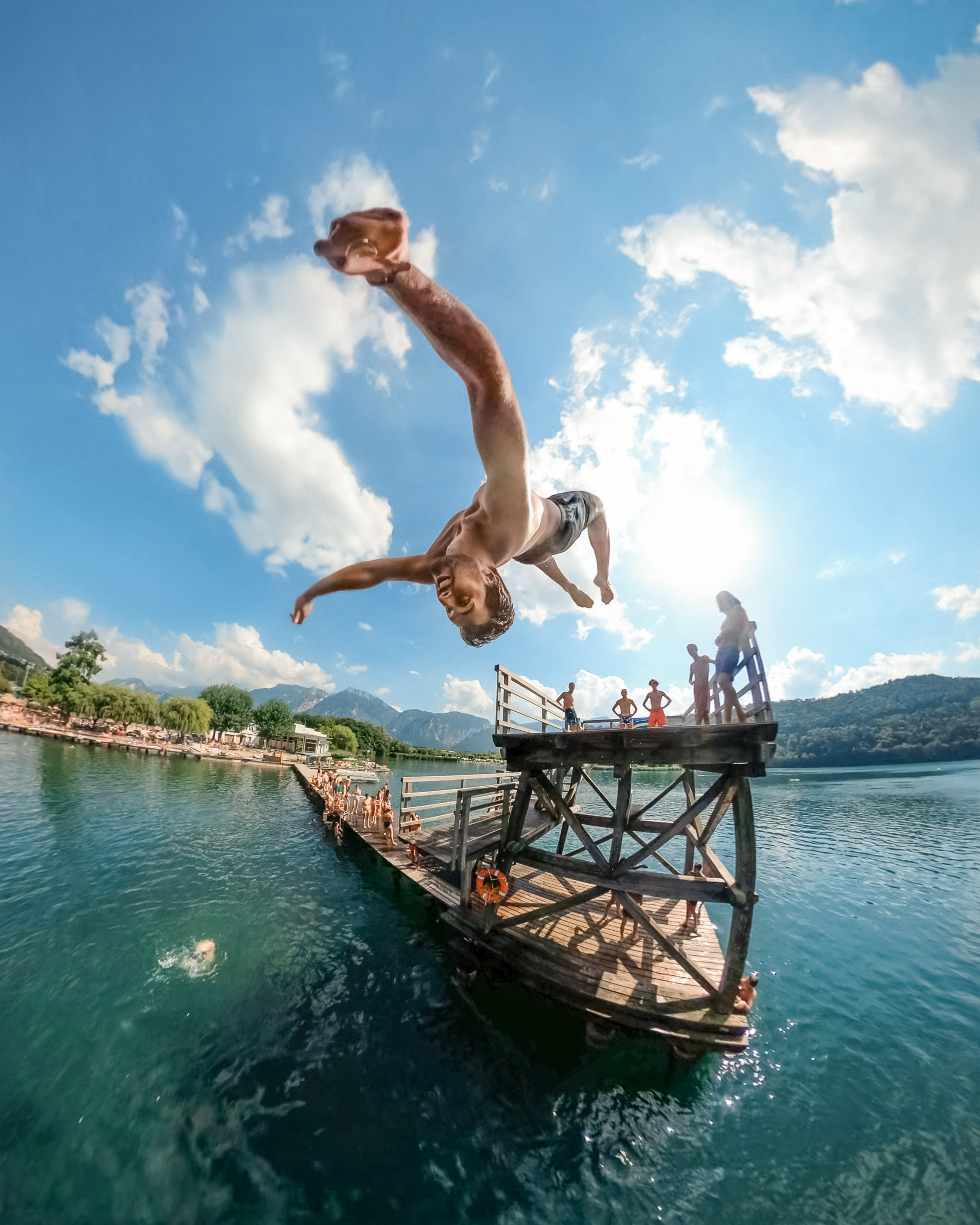 Mann springt von Holzkonstruktion in See. Blaues Wasser, sonniger Himmel, Zuschauer.
