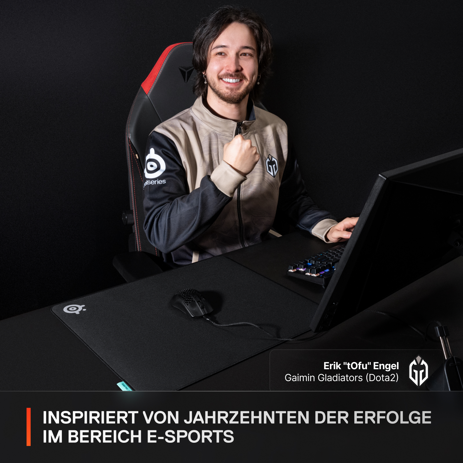 Gamer lächelt, Faust oben, am Schreibtisch mit Maus, Tastatur, Monitor.