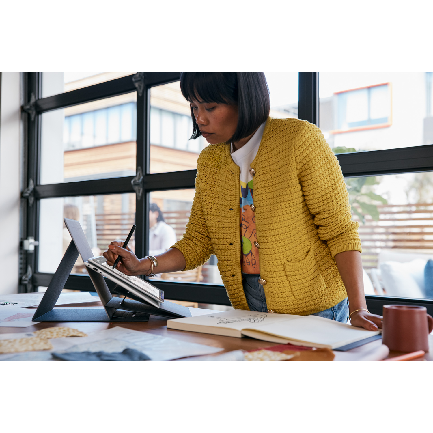 A woman working on a laptop, standing, and using a pen. There's a notebook, cup, and other items on the table.