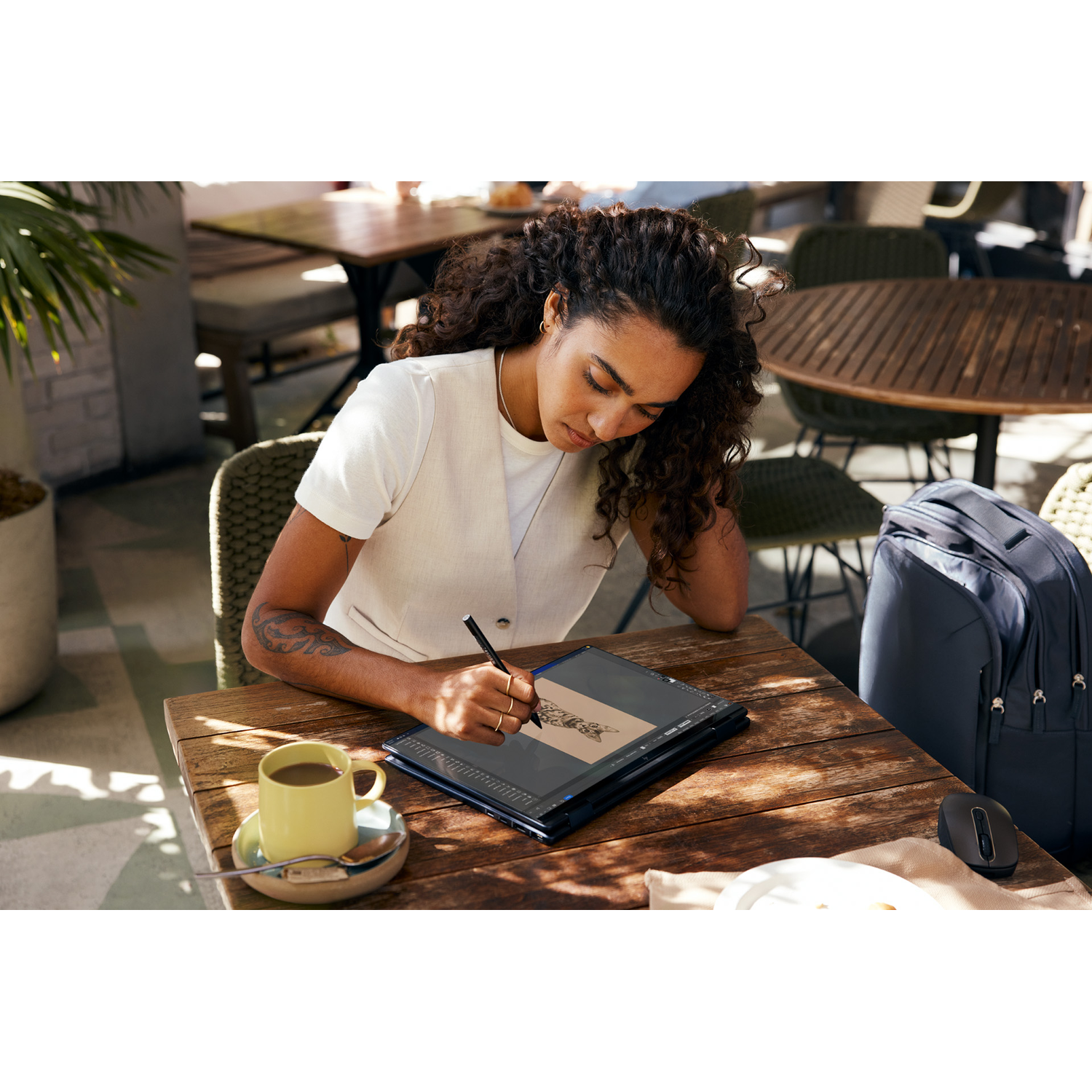 A woman is drawing on a tablet with a stylus while seated at a table, cafe setting, sunny day.
