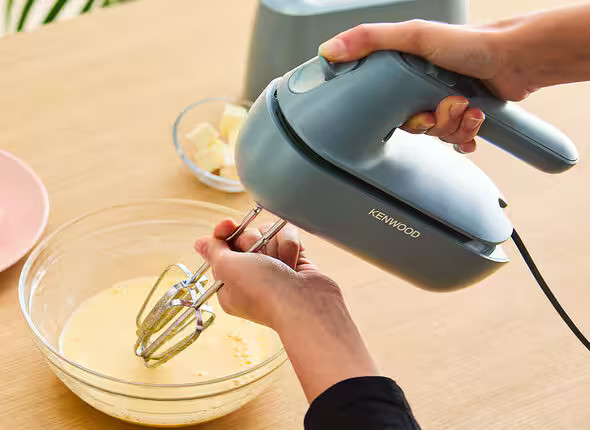 Dłonie osoby używającej szarego miksera ręcznego Kenwood do mieszania składników w szklanej misce.