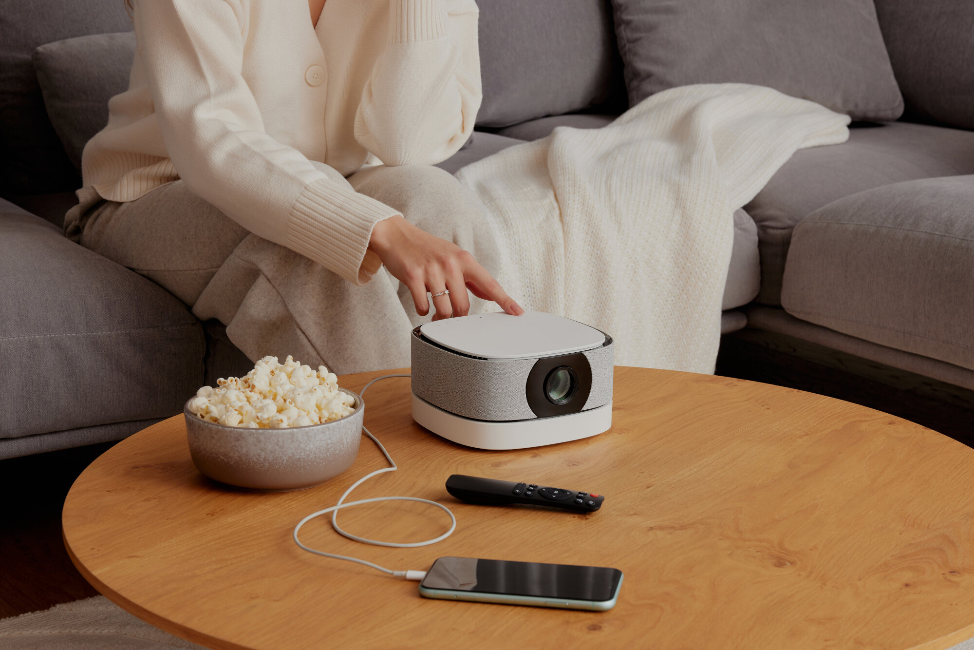 Osoba wskazuje na projektor na stole z popcornem, pilotem i telefonem.