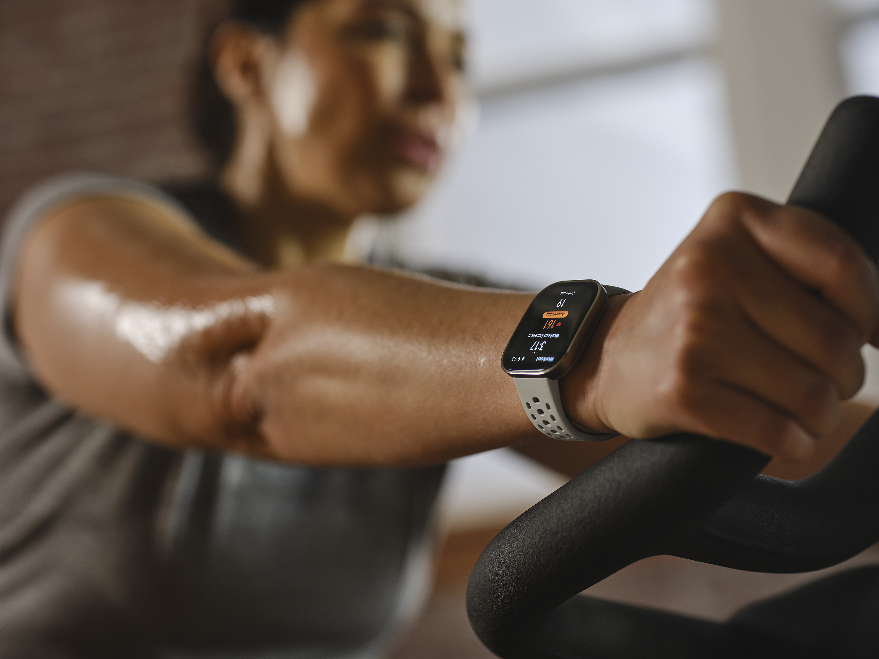 Egy személy karja okosórával, miközben szobabiciklin edz. Az óra fitnesz adatokat jelenít meg.