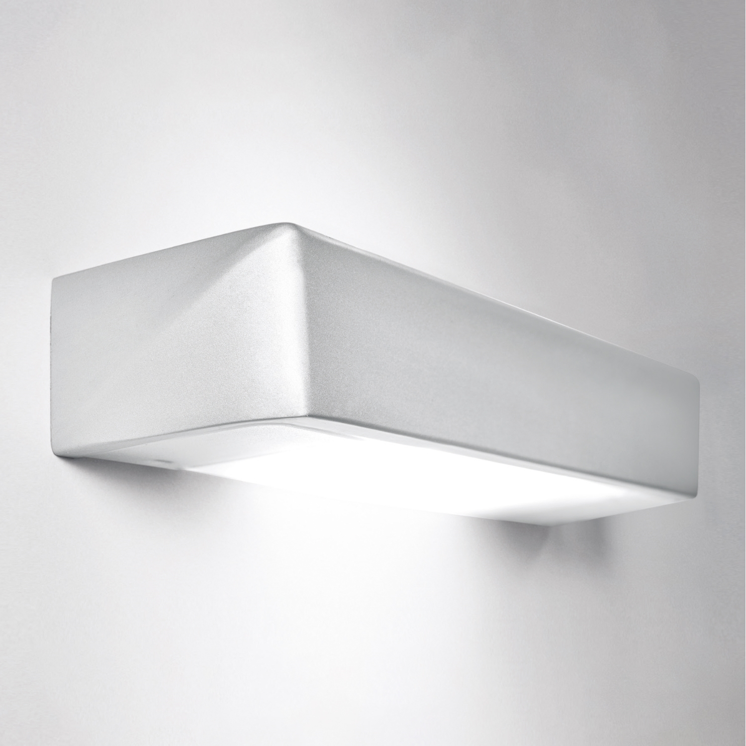 Lampada da parete rettangolare bianca, montata su un muro bianco. Emette una luce soffusa verso il basso.