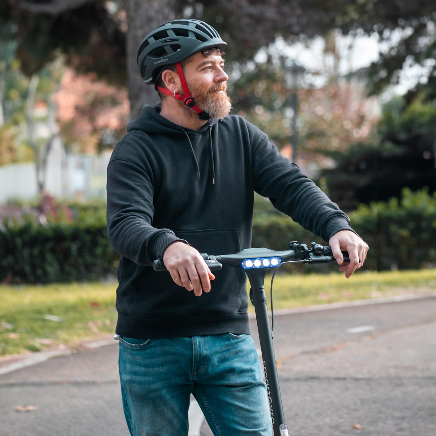 Hombre monta un patinete eléctrico negro en un camino. Lleva casco y sudadera con capucha.