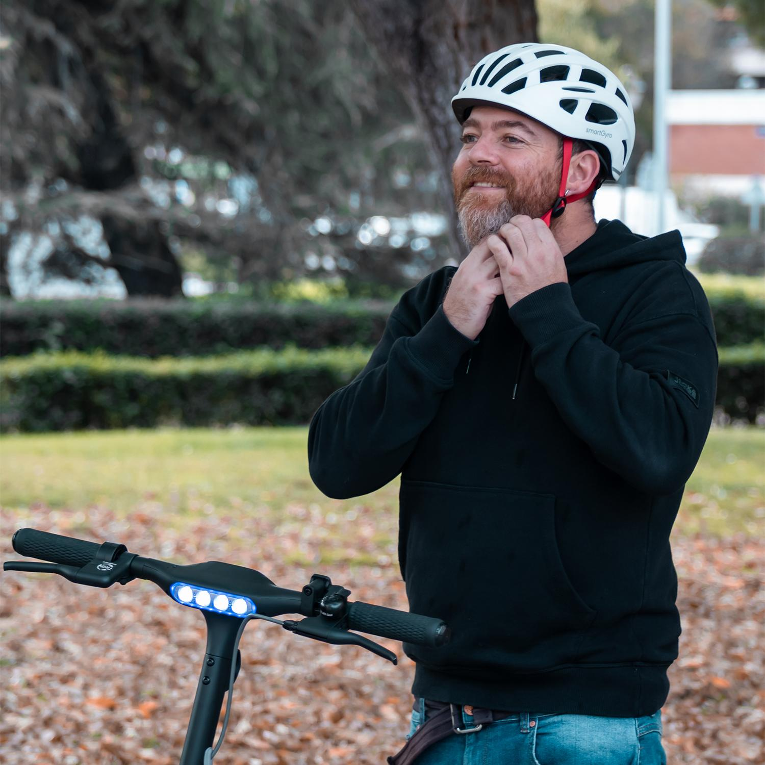 Un hombre ajusta un casco blanco. Está junto a un patinete eléctrico con barra de luz.