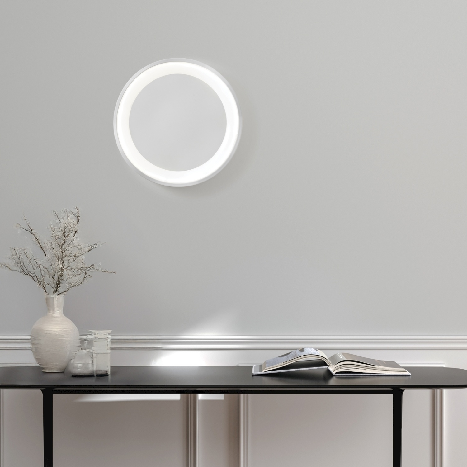Una lampada da parete bianca, rotonda e illuminata. Un vaso con rami bianchi e un libro su un tavolo.