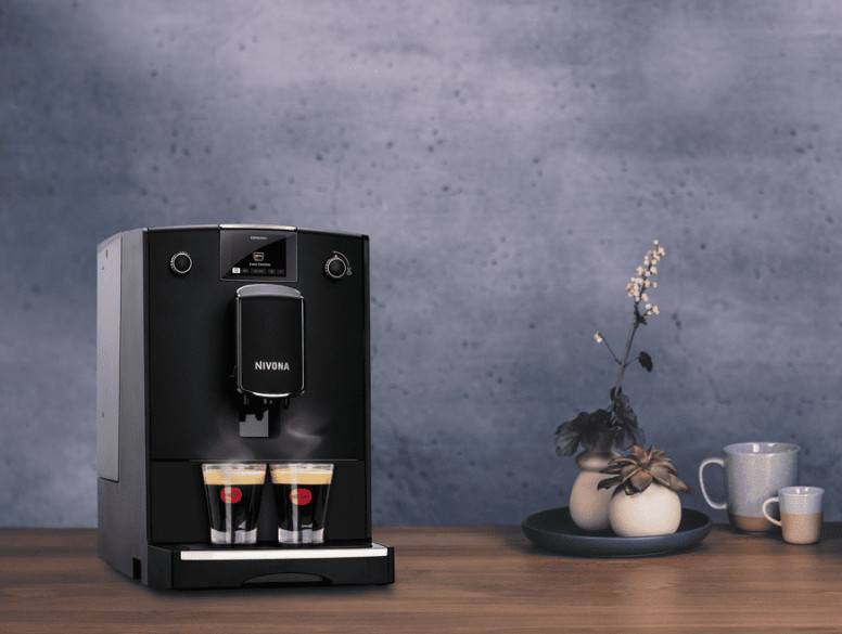 Czarny ekspres do kawy z dwoma szklankami wypełnionymi kawą. Tłem jest szara ściana i roślina.