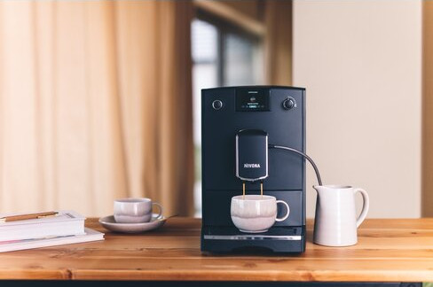 Czarny ekspres do kawy z filiżanką i dzbankiem na drewnianym stole. Zasłony w tle.