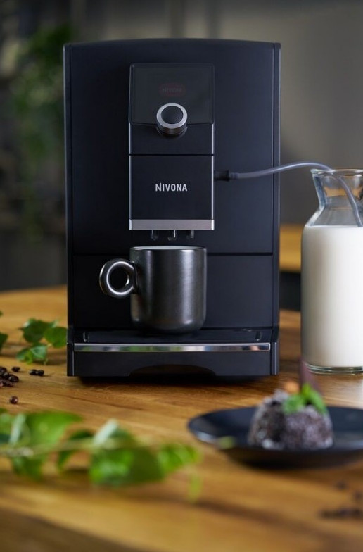 Czarny ekspres do kawy Nivona z filiżanką i dzbankiem na mleko na drewnianym stole. Małe ciasto na talerzu.