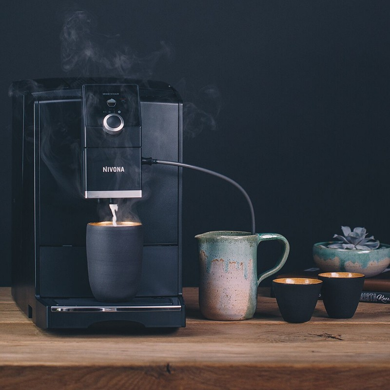 Ekspres do kawy nalewający do filiżanki. Kubek, filiżanki i roślina na drewnianym stole. Ciemne tło.