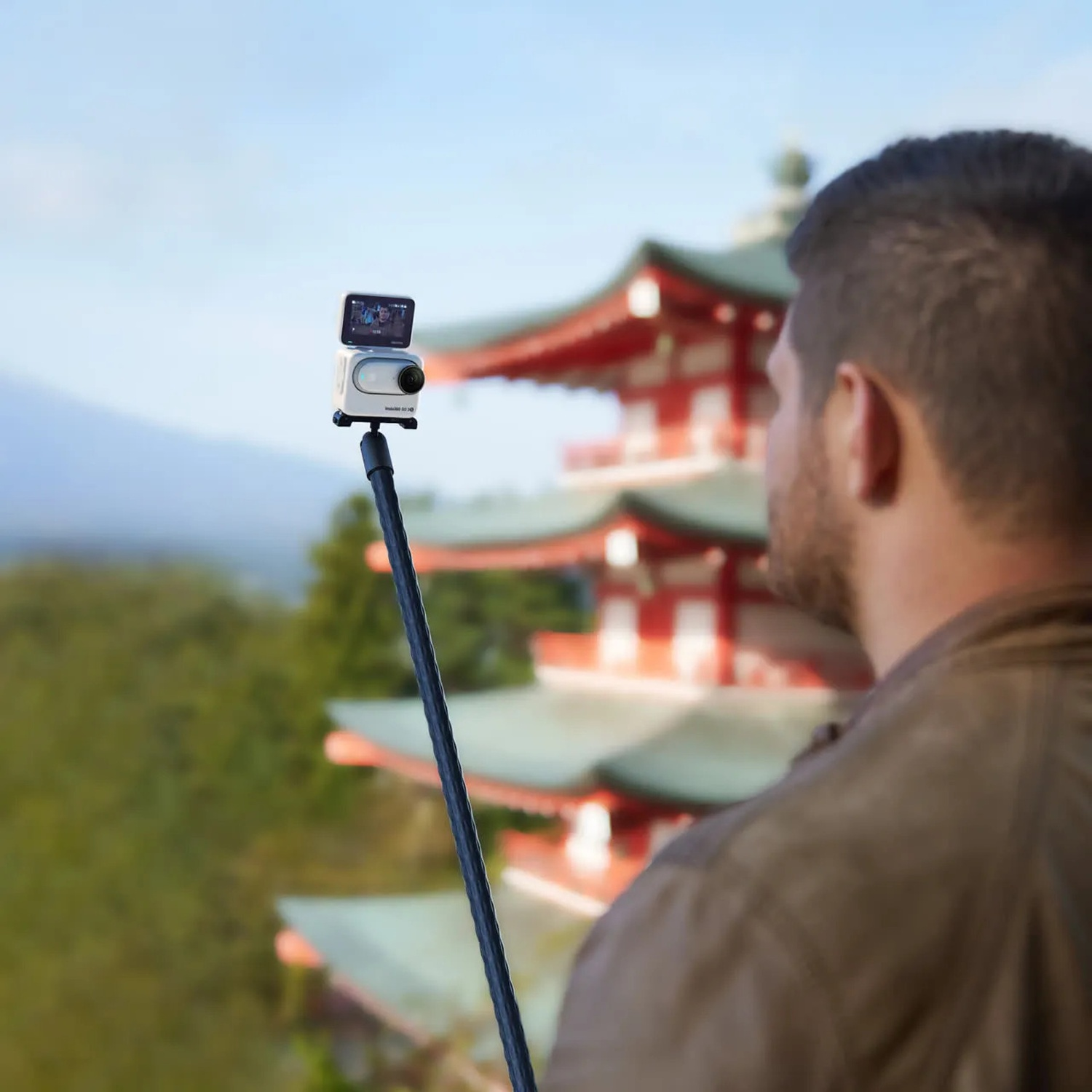 Un uomo tiene una fotocamera su un bastone selfie, con un tempio giapponese sullo sfondo.