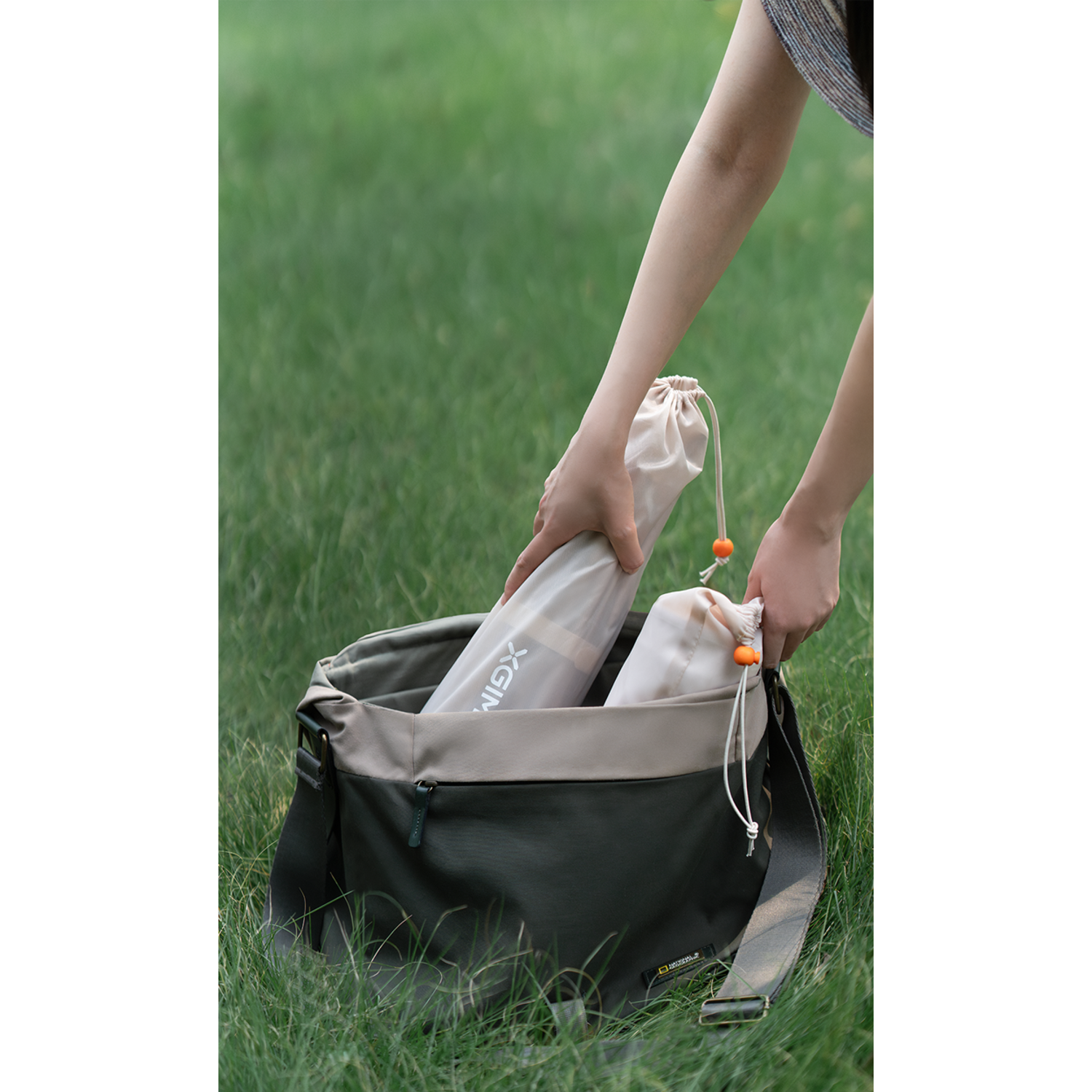Mains mettant des objets dans un sac, avec un sac vert-beige sur l'herbe.