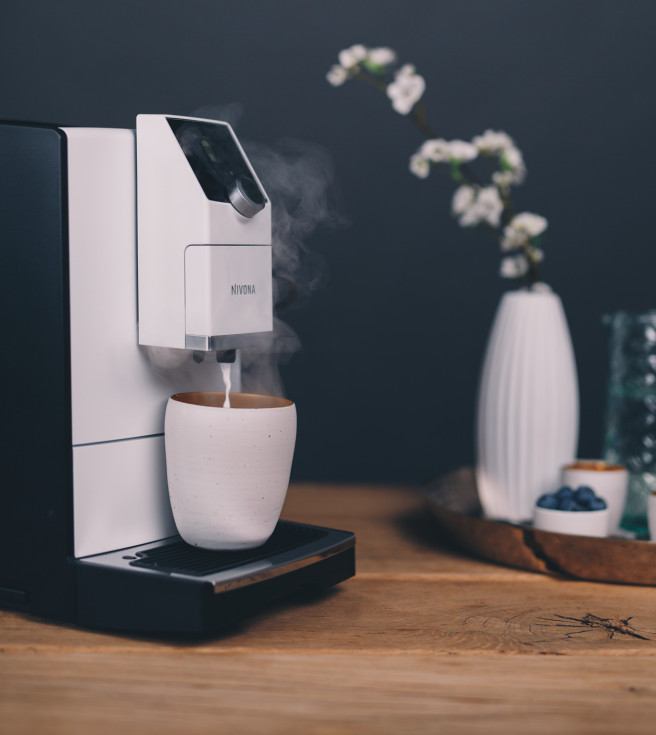 Biało-czarny ekspres do kawy z parą, filiżanka i wazon z kwiatami.