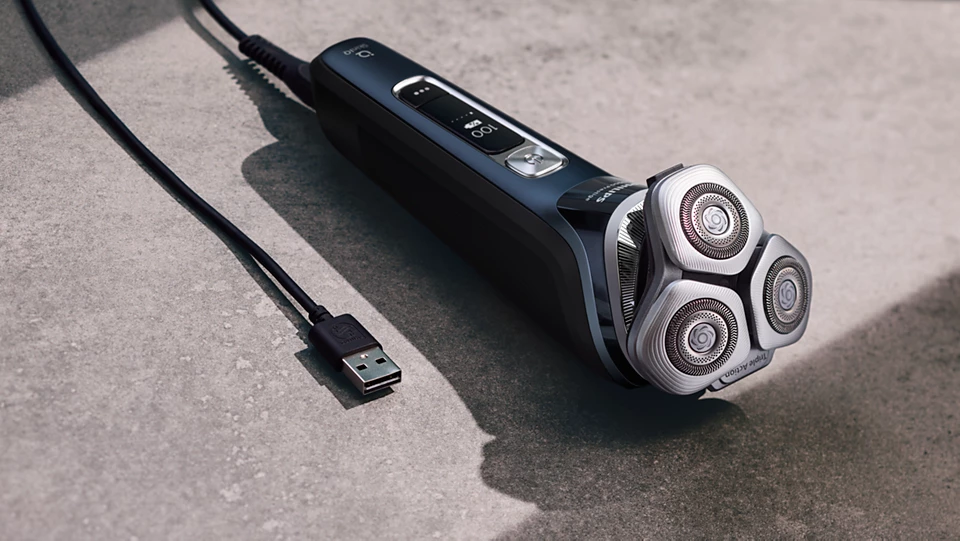 Schwarz-silberner Elektrorasierer. Drei rotierende Köpfe. USB-Kabel. Graue Oberfläche.