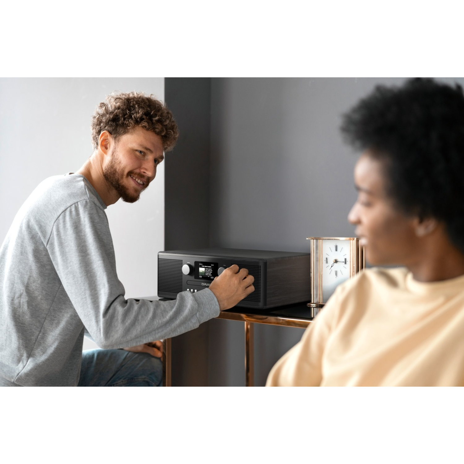 Ein Mann stellt die Lautstärke eines Holzradios ein und schaut eine Frau an. In der Nähe steht eine Uhr.