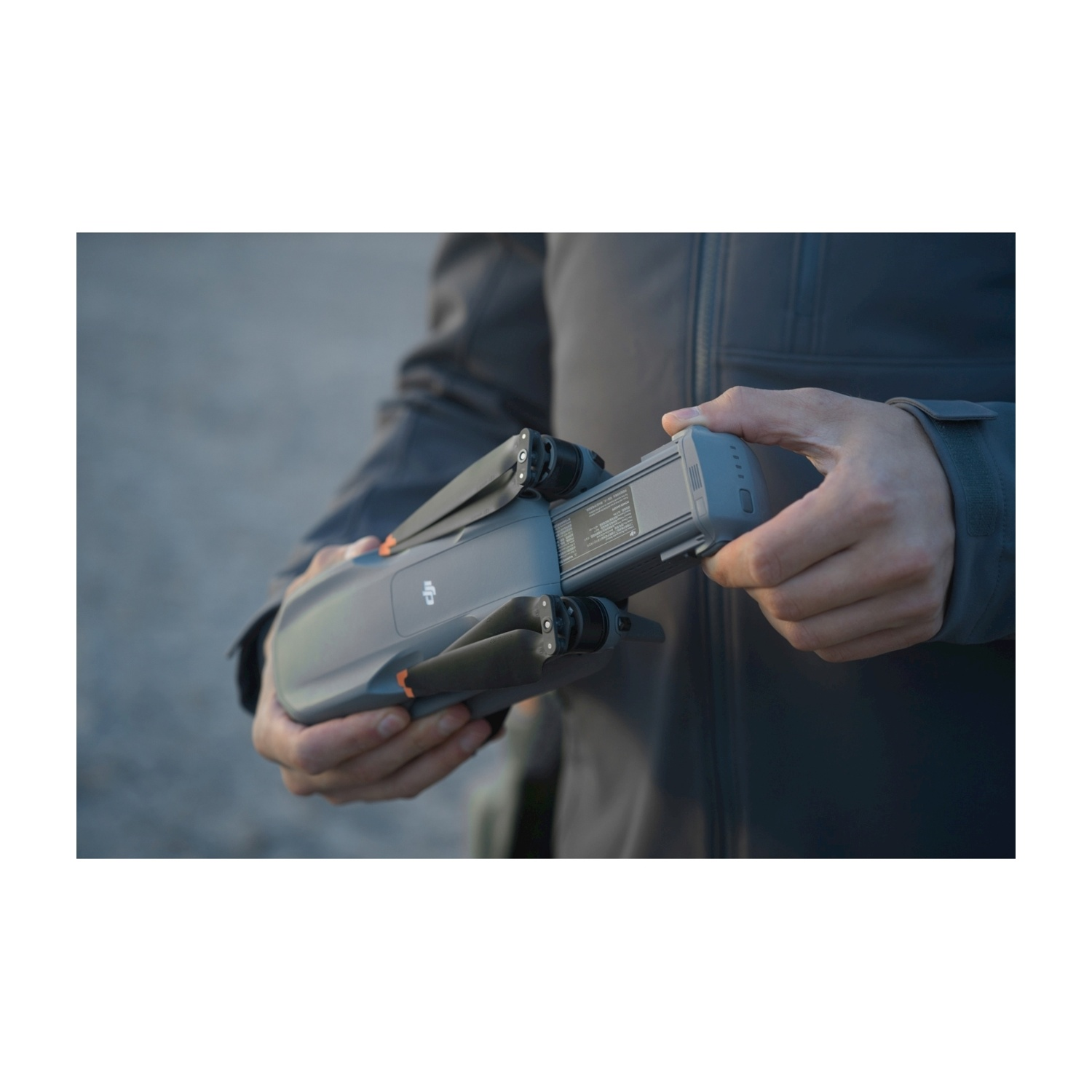 Una persona tiene in mano un drone grigio, inserendo una batteria. Lo sfondo è sfocato.