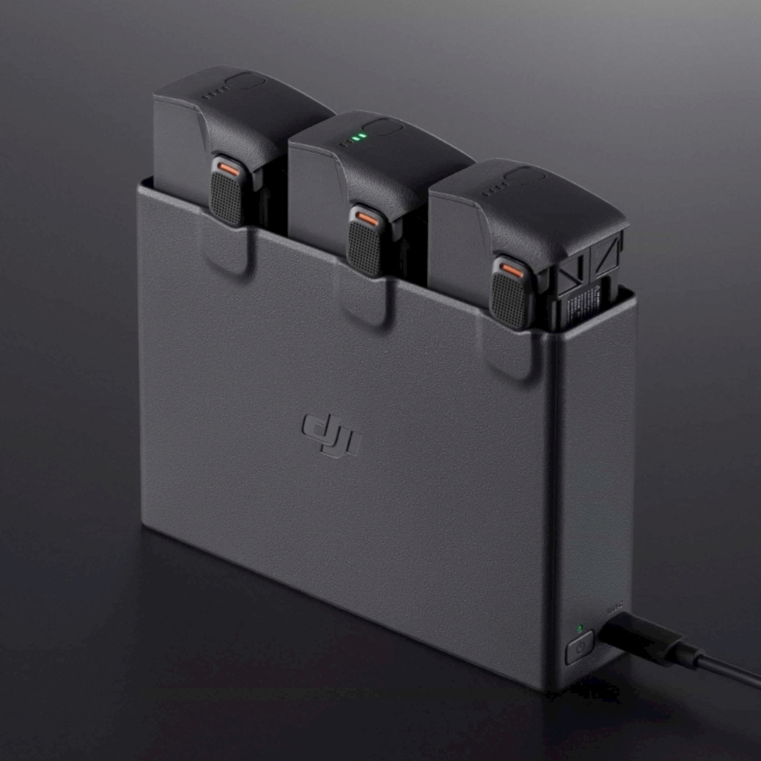 Una custodia di ricarica rettangolare grigia con tre batterie nere inserite. Un cavo di ricarica nero è collegato.