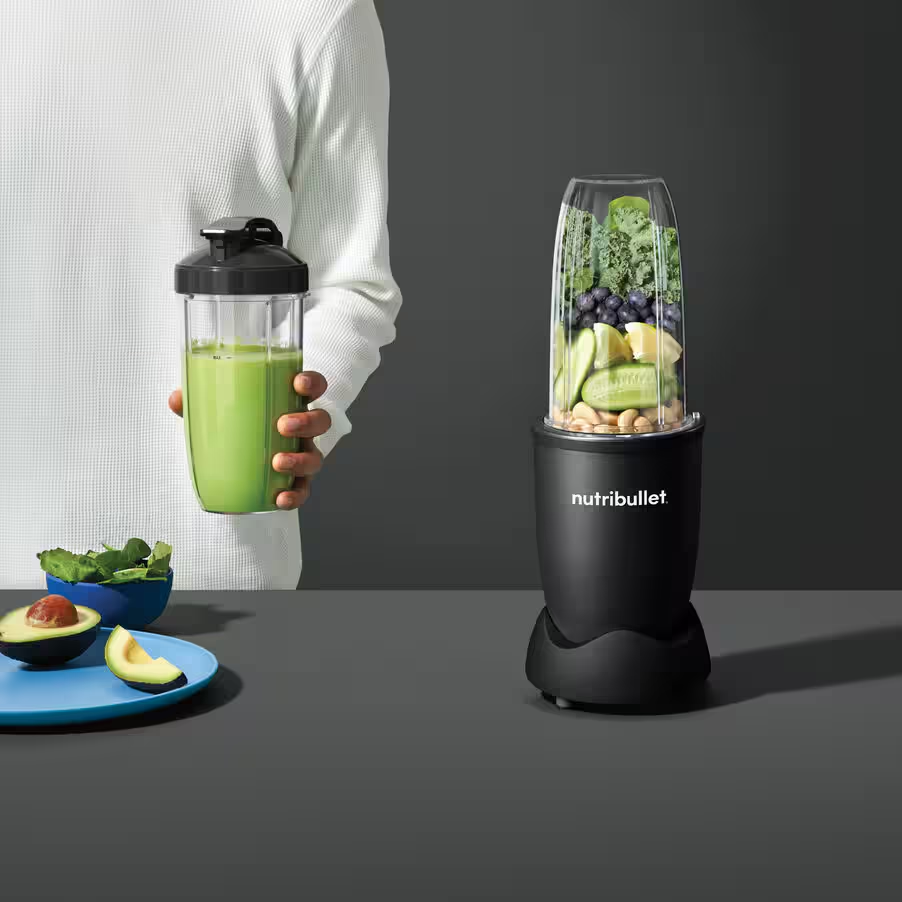 Personne tenant un smoothie vert dans un gobelet près d'un mixeur.