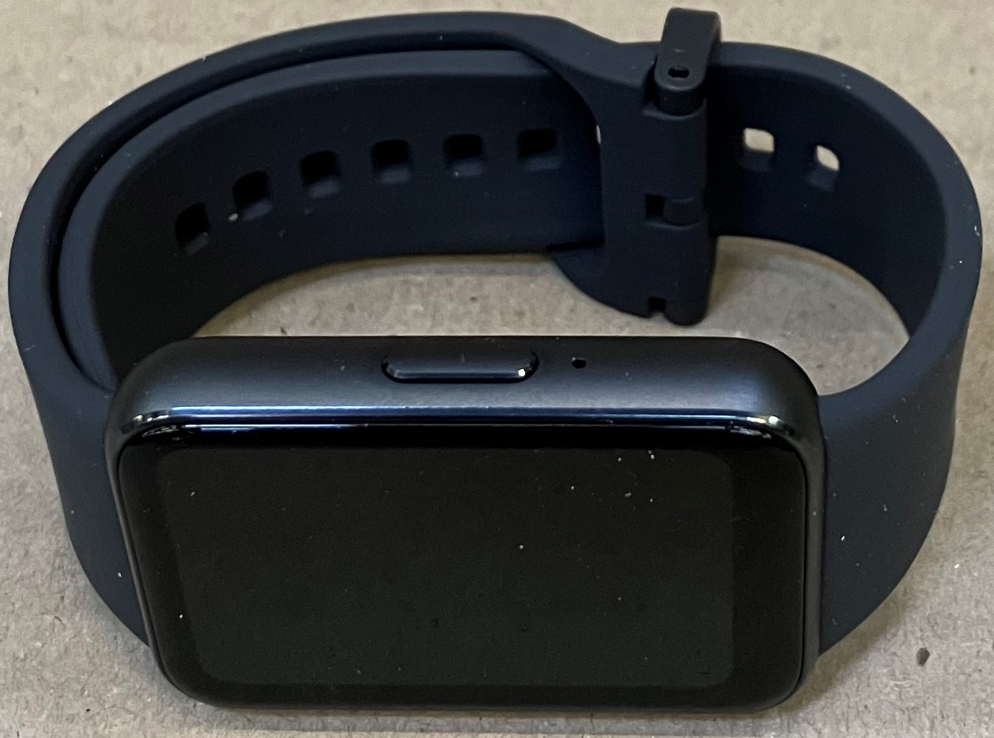 Czarny smartwatch z prostokątnym ekranem i czarnym paskiem. Zegarek leży na powierzchni.
