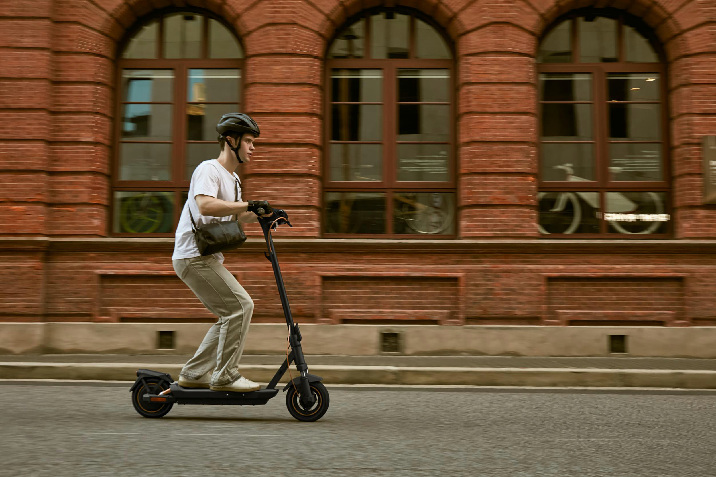 Ein Mann fährt mit einem E-Scooter auf einer Stadtstraße. Backsteingebäude im Hintergrund.