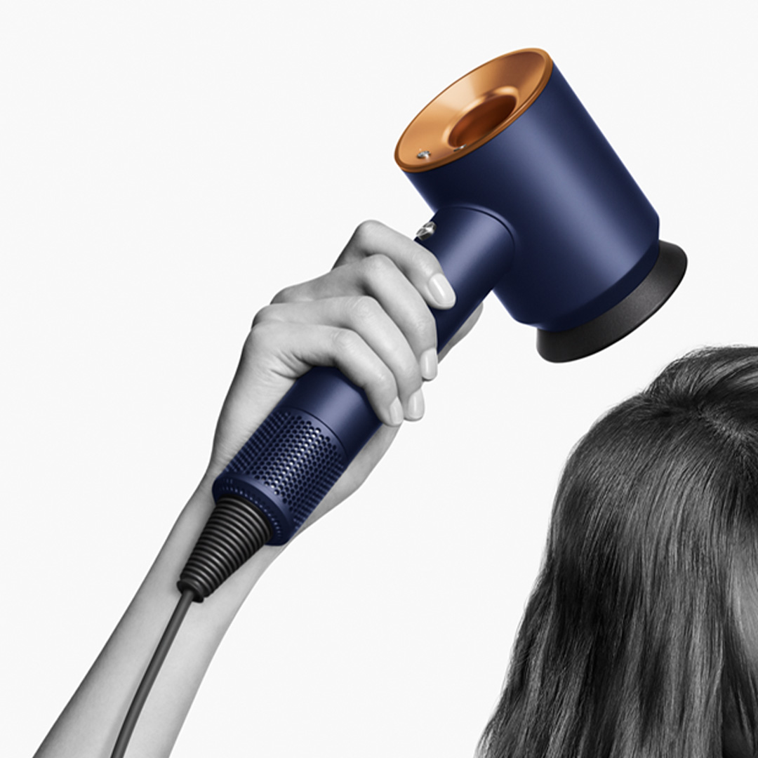 Osoba trzymająca niebiesko-złotą suszarkę do włosów, susząca włosy. Tło jest jasnoszare.