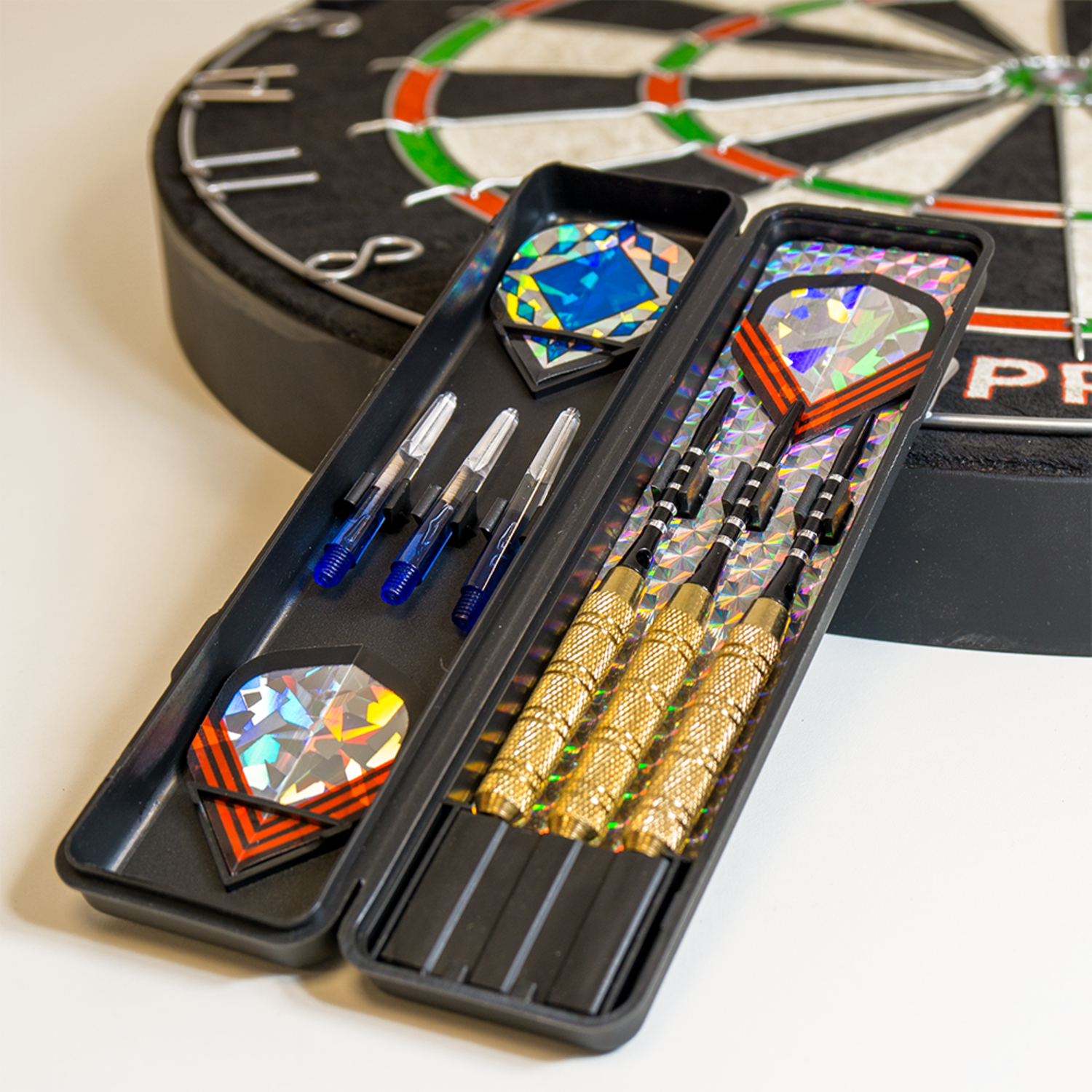 Darts en accessoires in een zwarte koffer, met een dartbord op de achtergrond.
