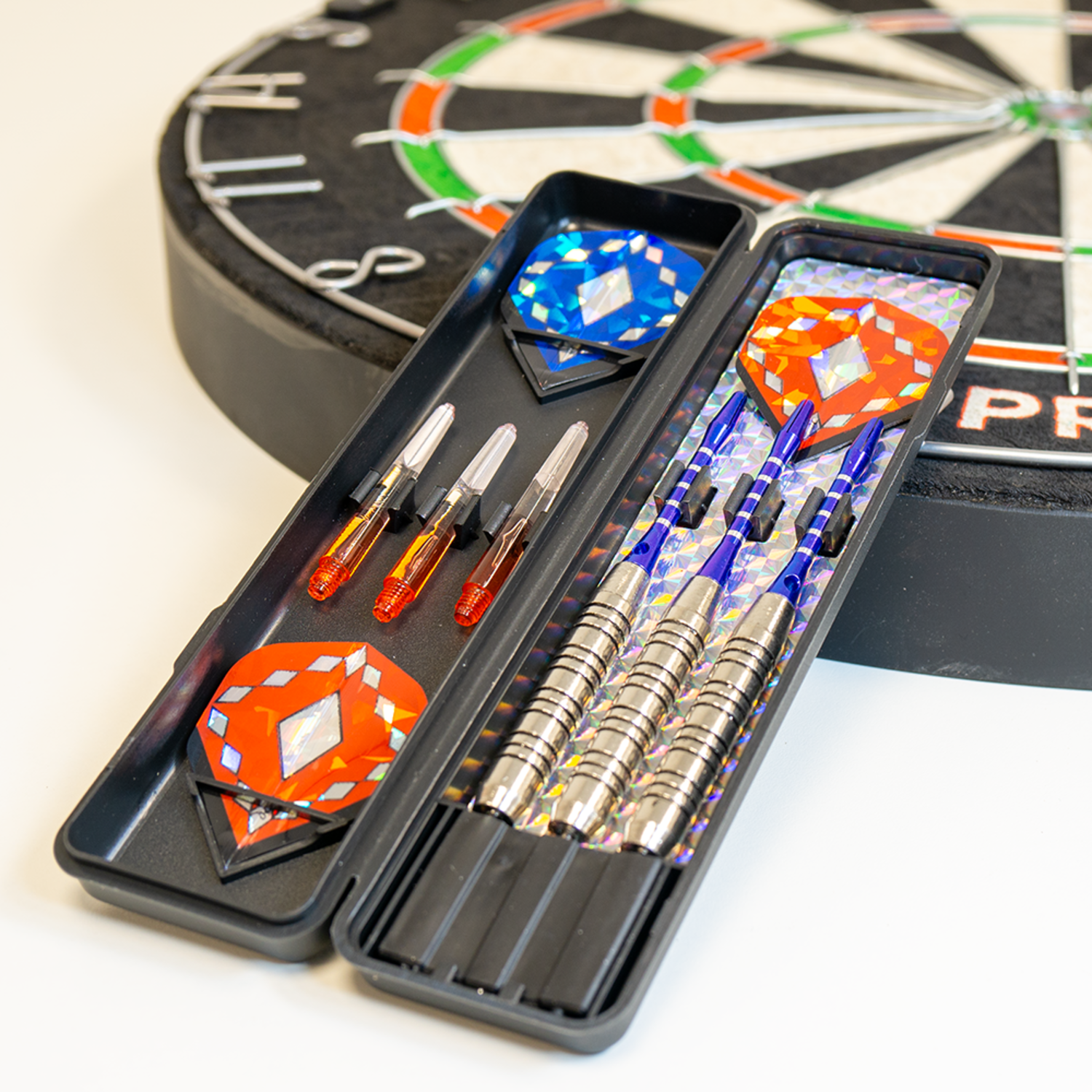 Dartbord en darts in een etui. Darts hebben blauwe en zilveren schachten, oranje flights. Etui is open, toont items. Witte achtergrond.