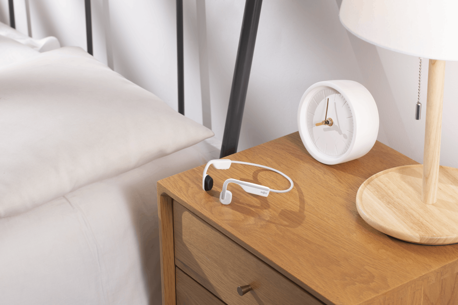 Auriculares blancos en una mesita de noche de madera junto a un reloj y una lámpara blancos con una cama blanca.