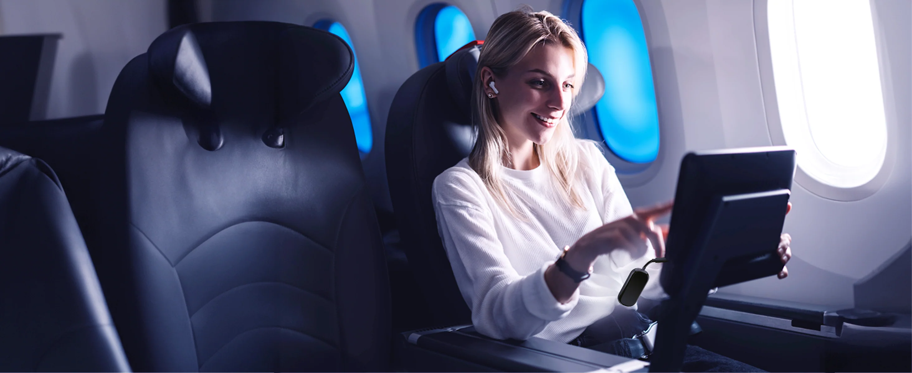 Une femme dans un avion utilise une tablette. Elle sourit et porte des écouteurs. Intérieur de l'avion.