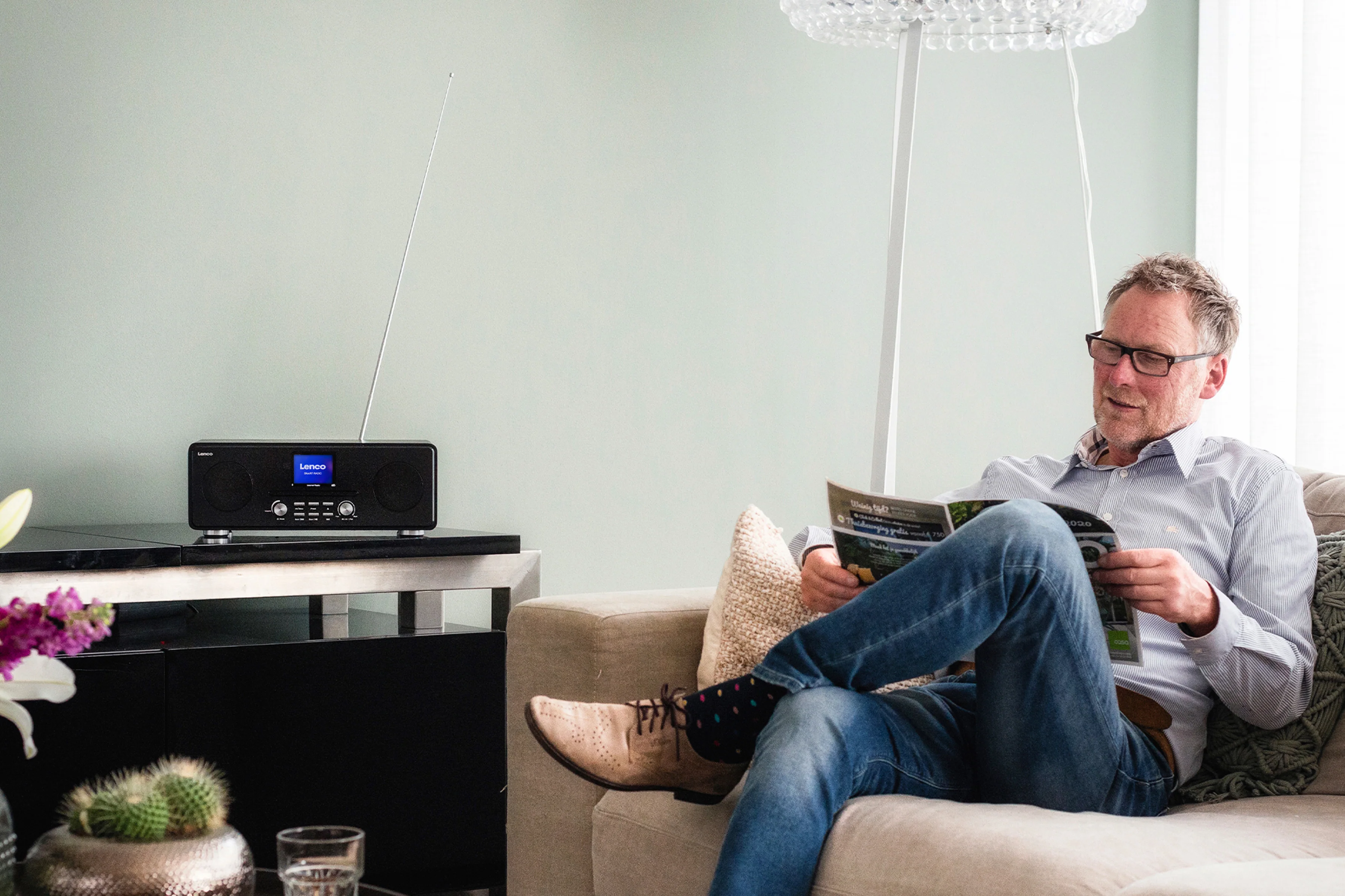 Homme lisant un magazine sur un canapé, radio sur une table. Lampe et cactus visibles.