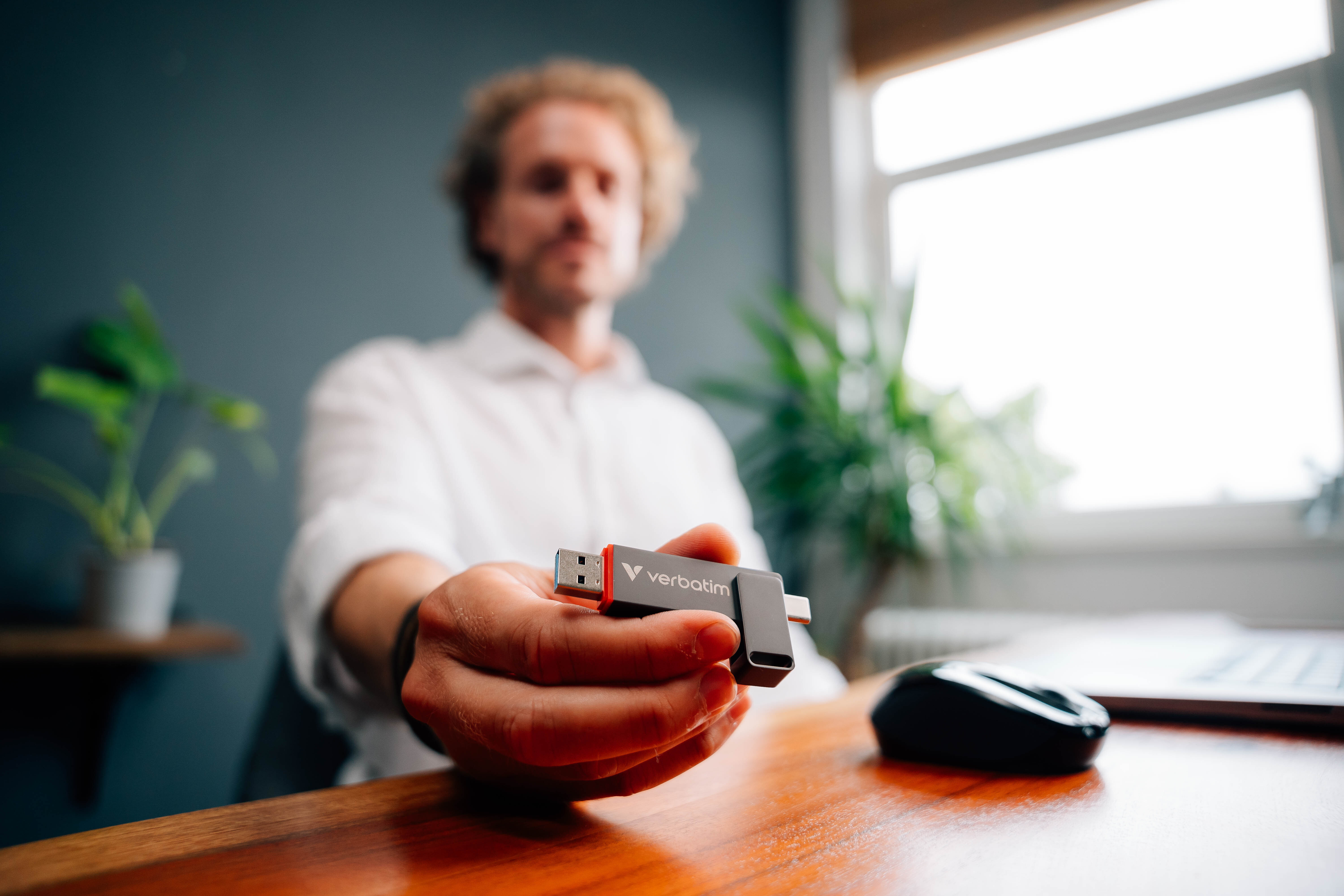 Ein Mann hält einen USB-Stick. Schreibtisch mit Pflanze, Maus und Fenster im Hintergrund.