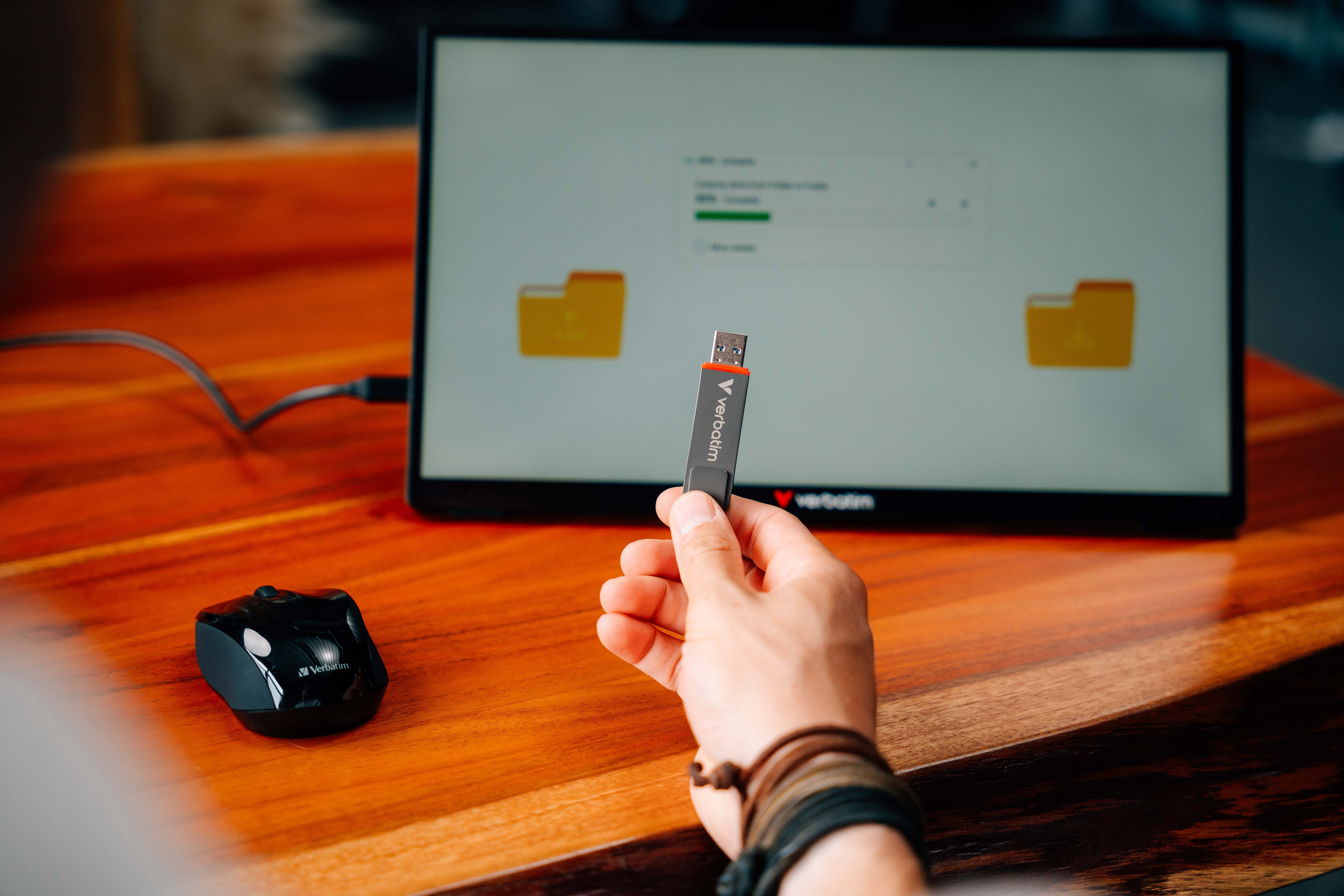 Person hält einen schwarzen Verbatim USB-Stick vor einem Computermonitor, der eine Dateiübertragung anzeigt.