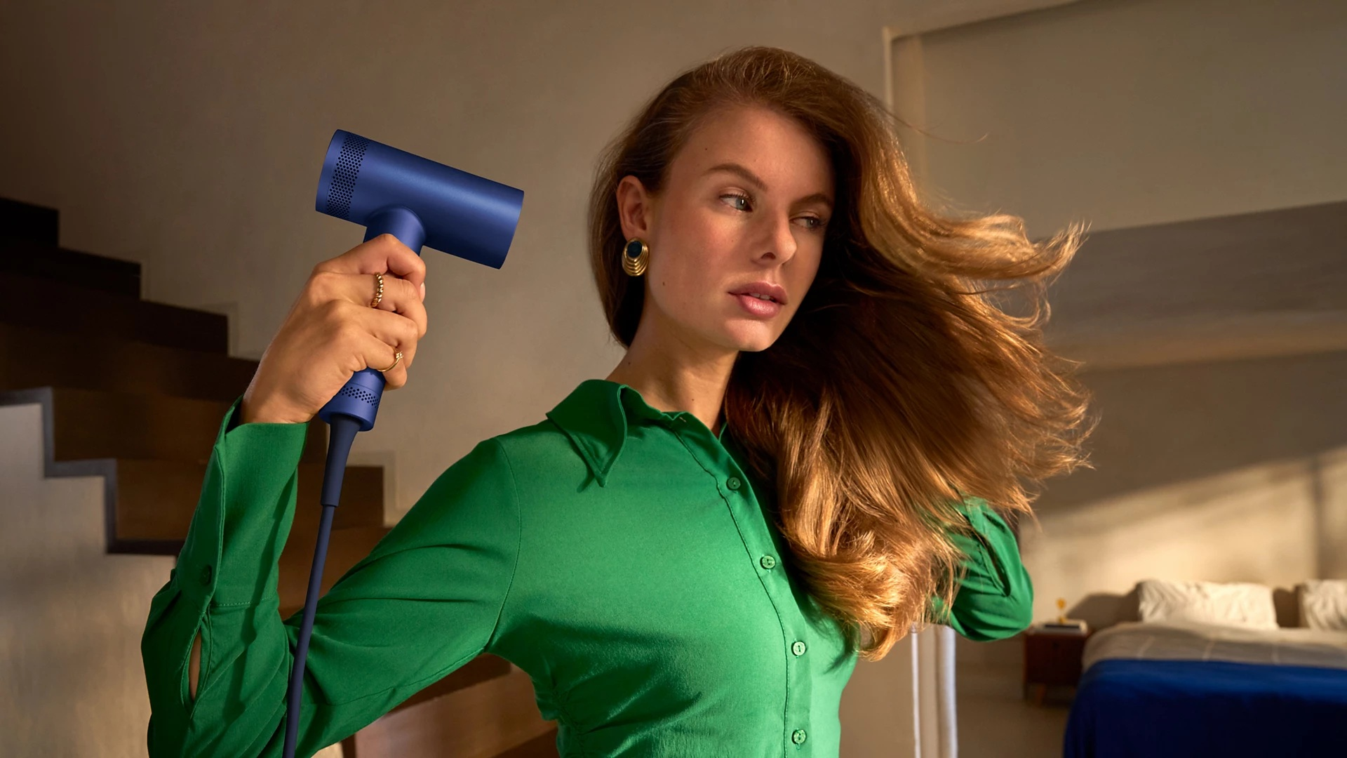 Kobieta suszy włosy niebieską suszarką. Tło to wnętrze domu.