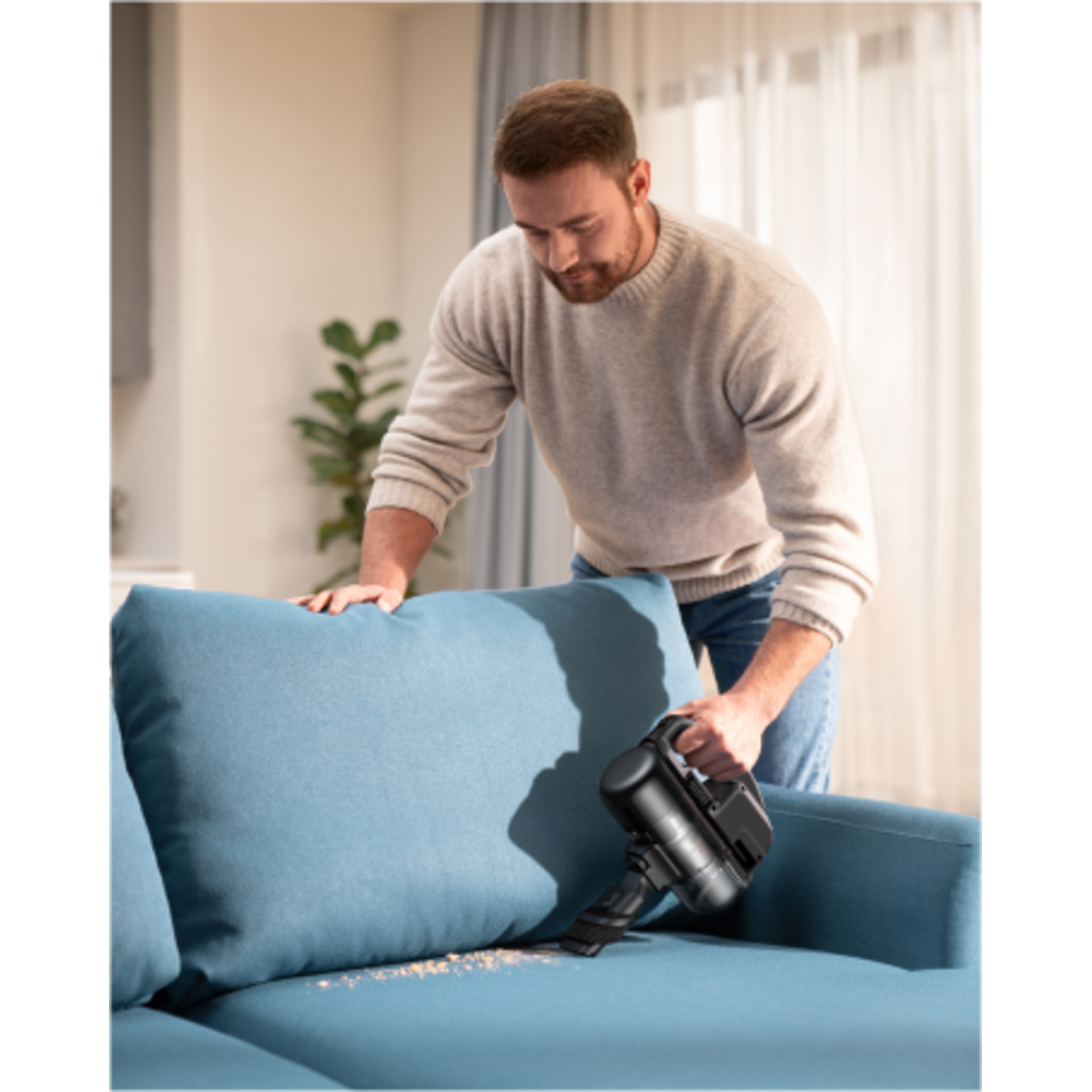 A man vacuums a blue sofa. Crumbs are visible on the cushion. He holds a black vacuum cleaner.