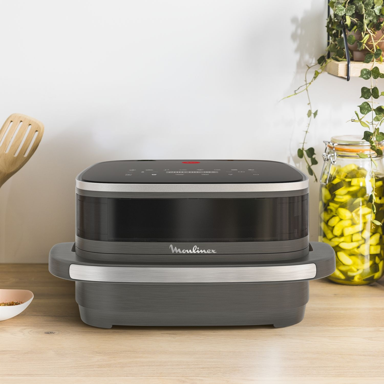 A black and silver Moulinex appliance sits on a wooden surface. A wooden spatula and a jar of pickles are also visible.