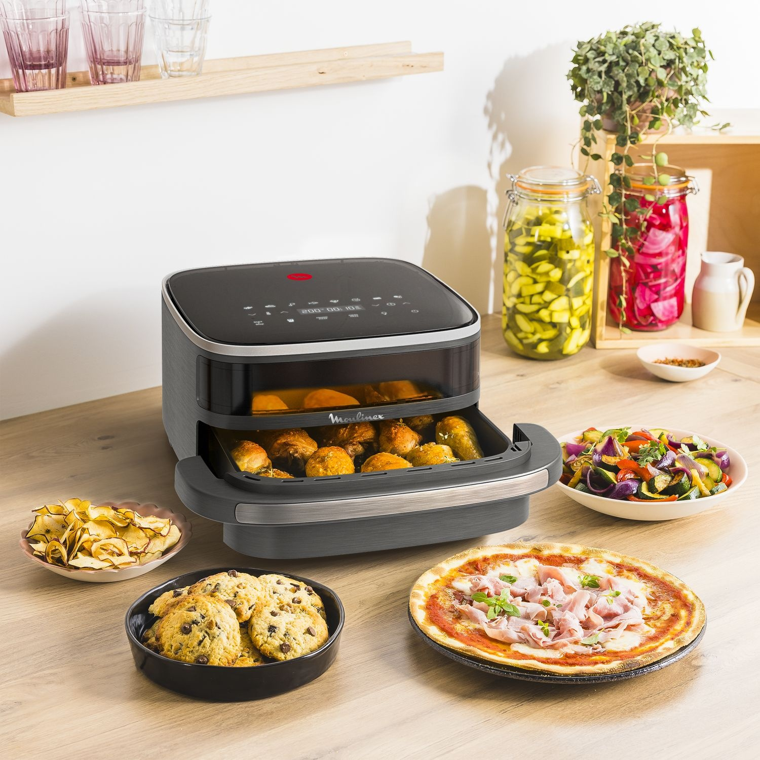 An air fryer with cooked food, pizza, cookies, chips, and vegetables. Jars of pickled items and glasses sit on a wooden shelf.