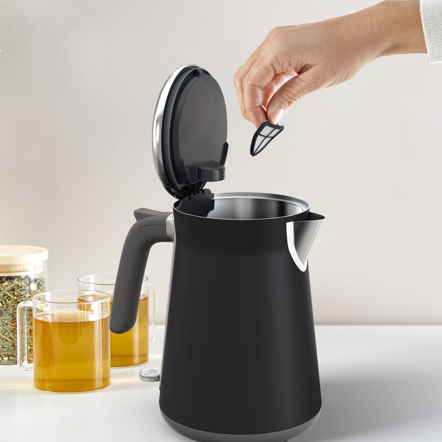 A hand removing a filter from a black kettle. Two tea glasses in the background.
