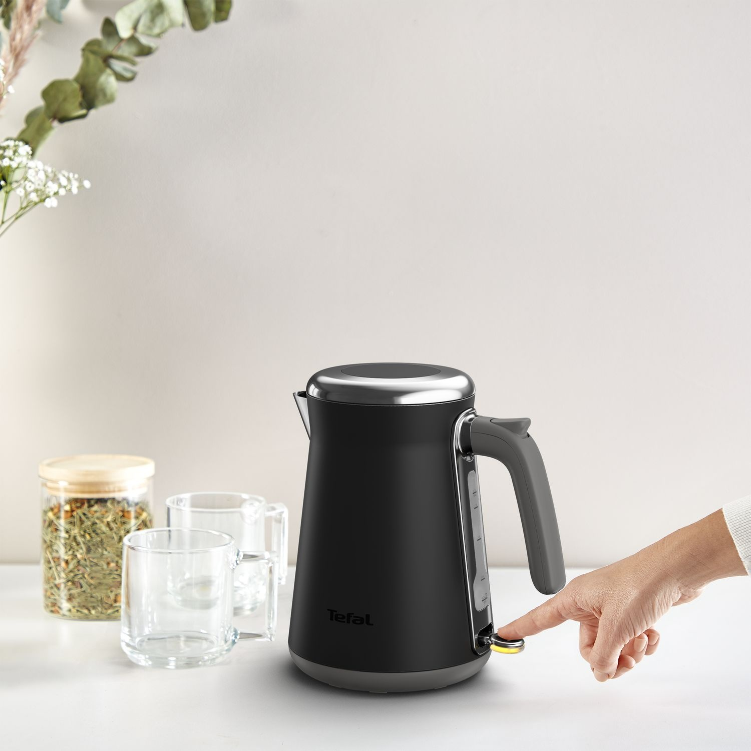 A black Tefal kettle, on a white surface. A hand is pressing the power button.