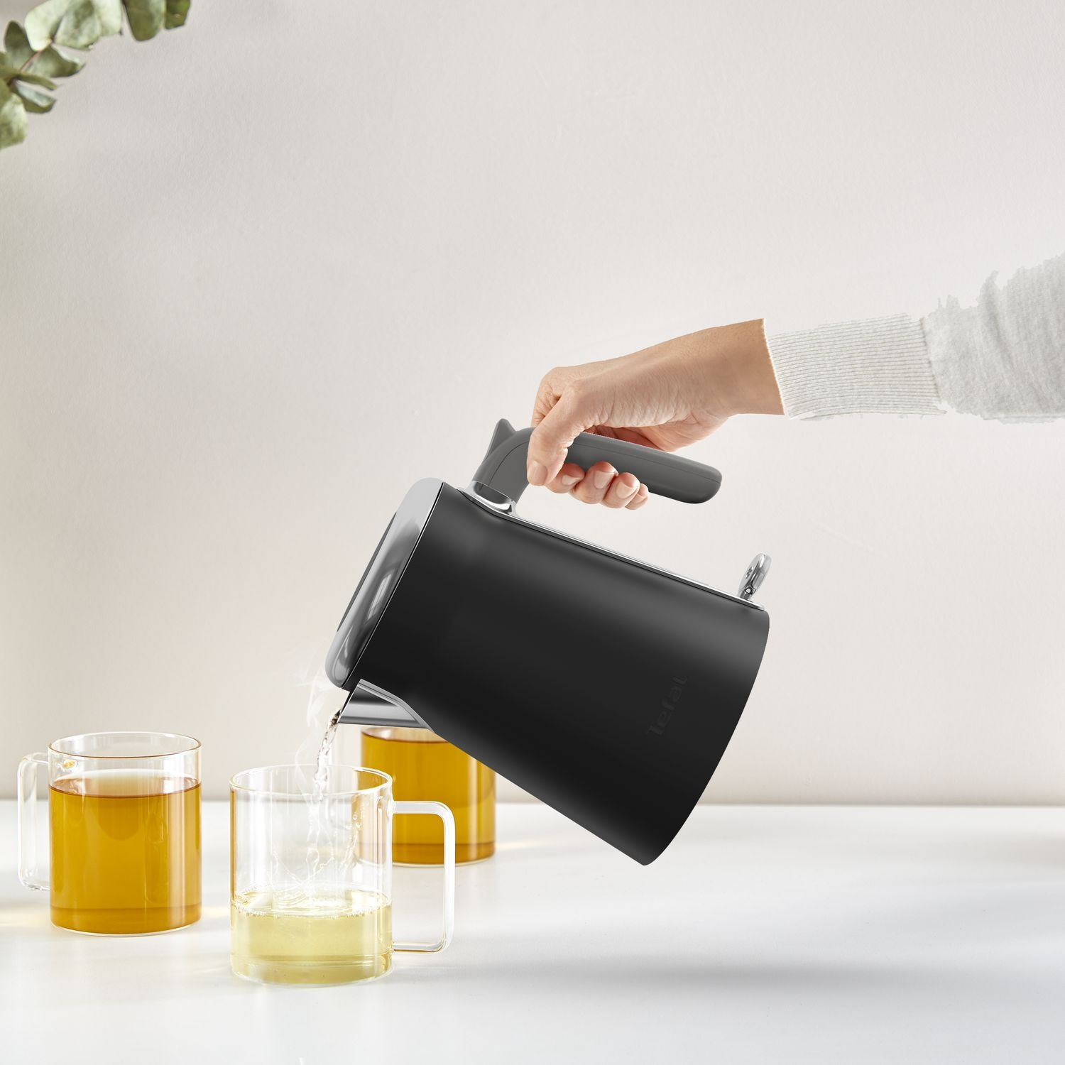 A person pouring water from a black kettle into a glass mug with tea.