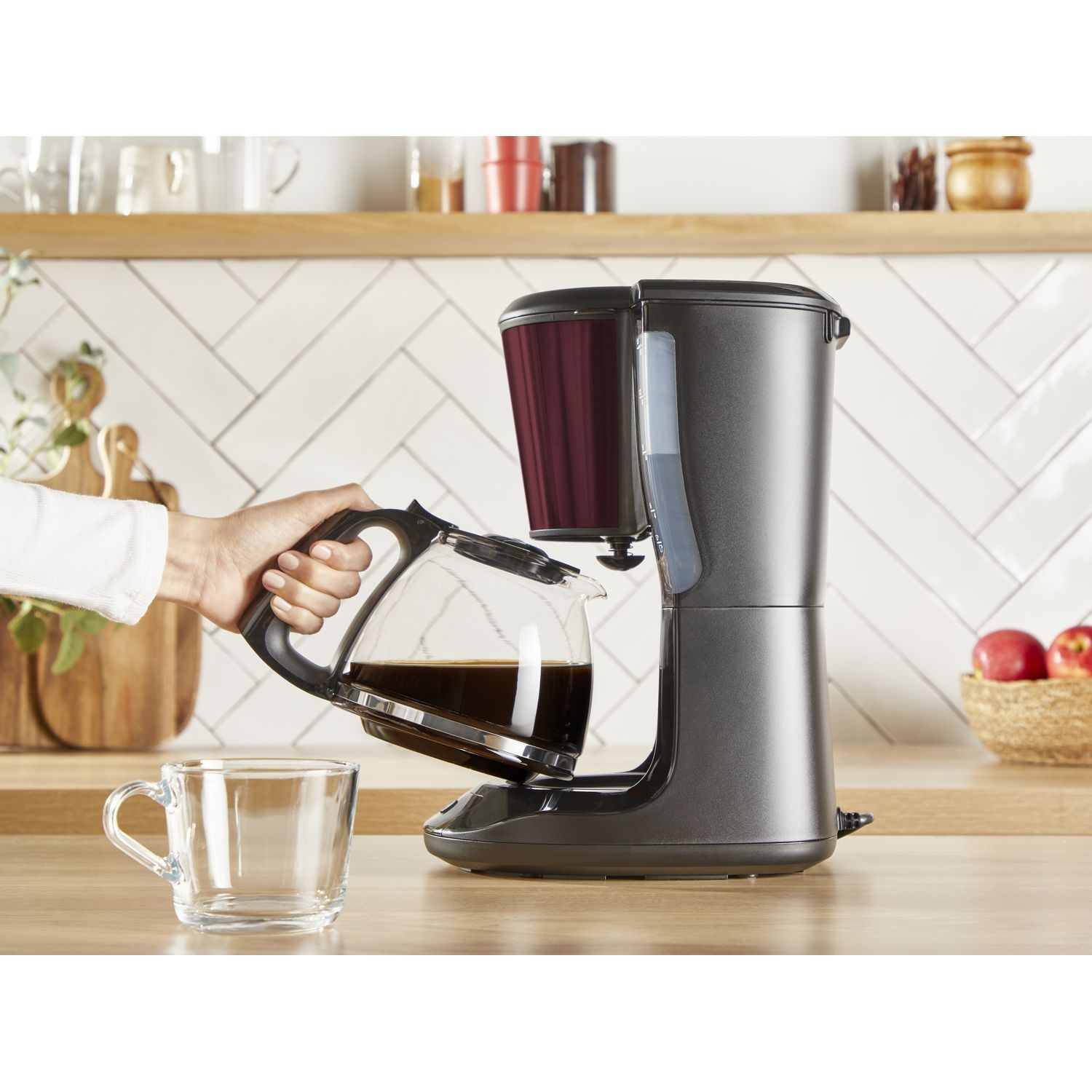 A hand pours coffee from a glass carafe into a clear glass mug on a wooden counter.