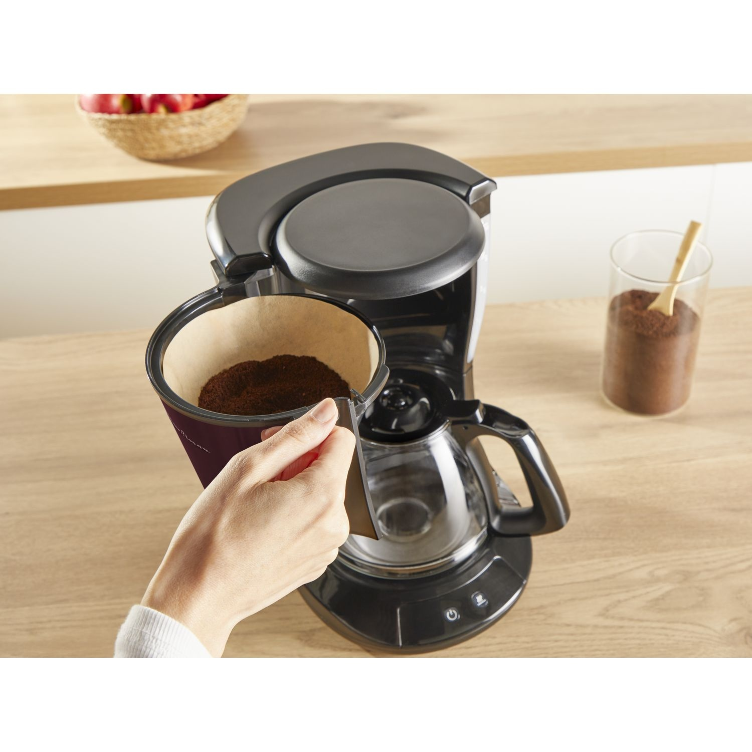 A hand places a coffee filter into a coffee maker. Coffee grounds are visible.