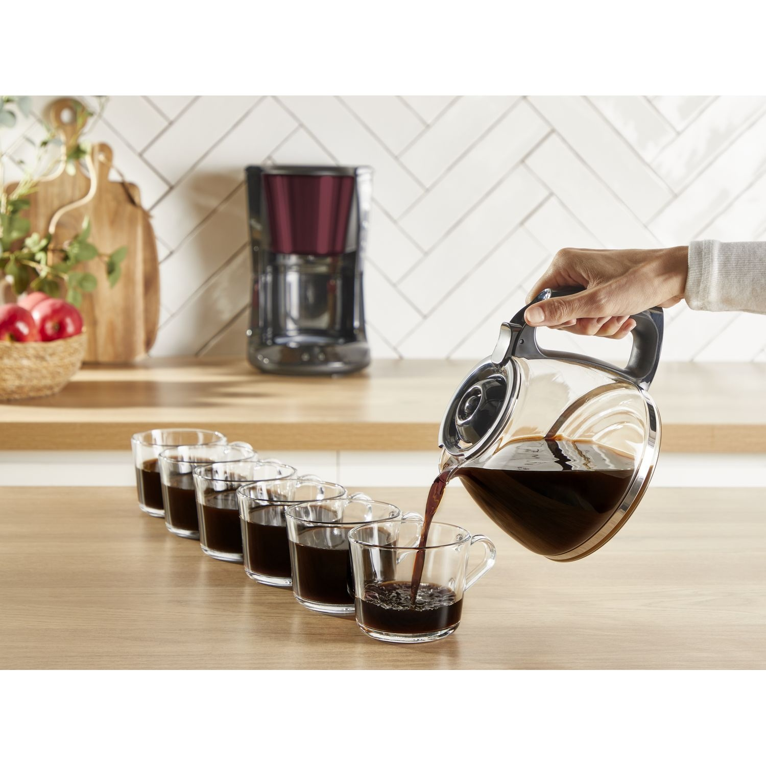A hand pours coffee from a glass carafe into a row of clear glass cups.