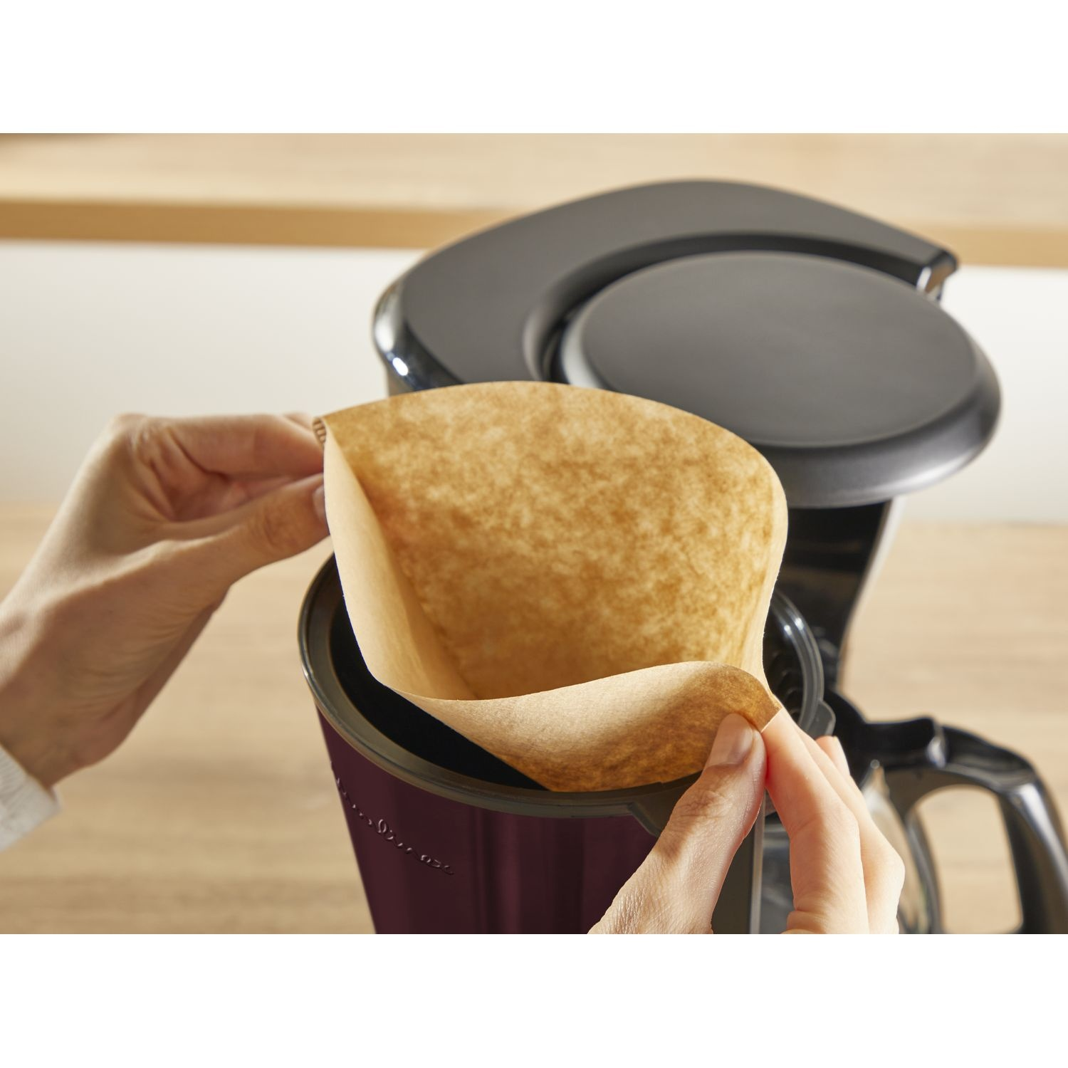 Hands insert a brown coffee filter into a coffee maker.