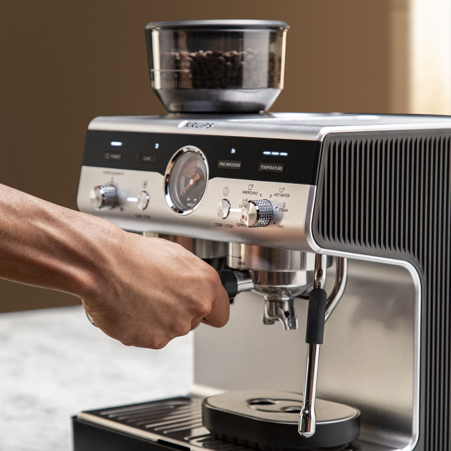 Une machine à expresso Krups argentée avec une main tenant un porte-filtre. Il y a une trémie à grains sur le dessus. La machine est sur marbre.