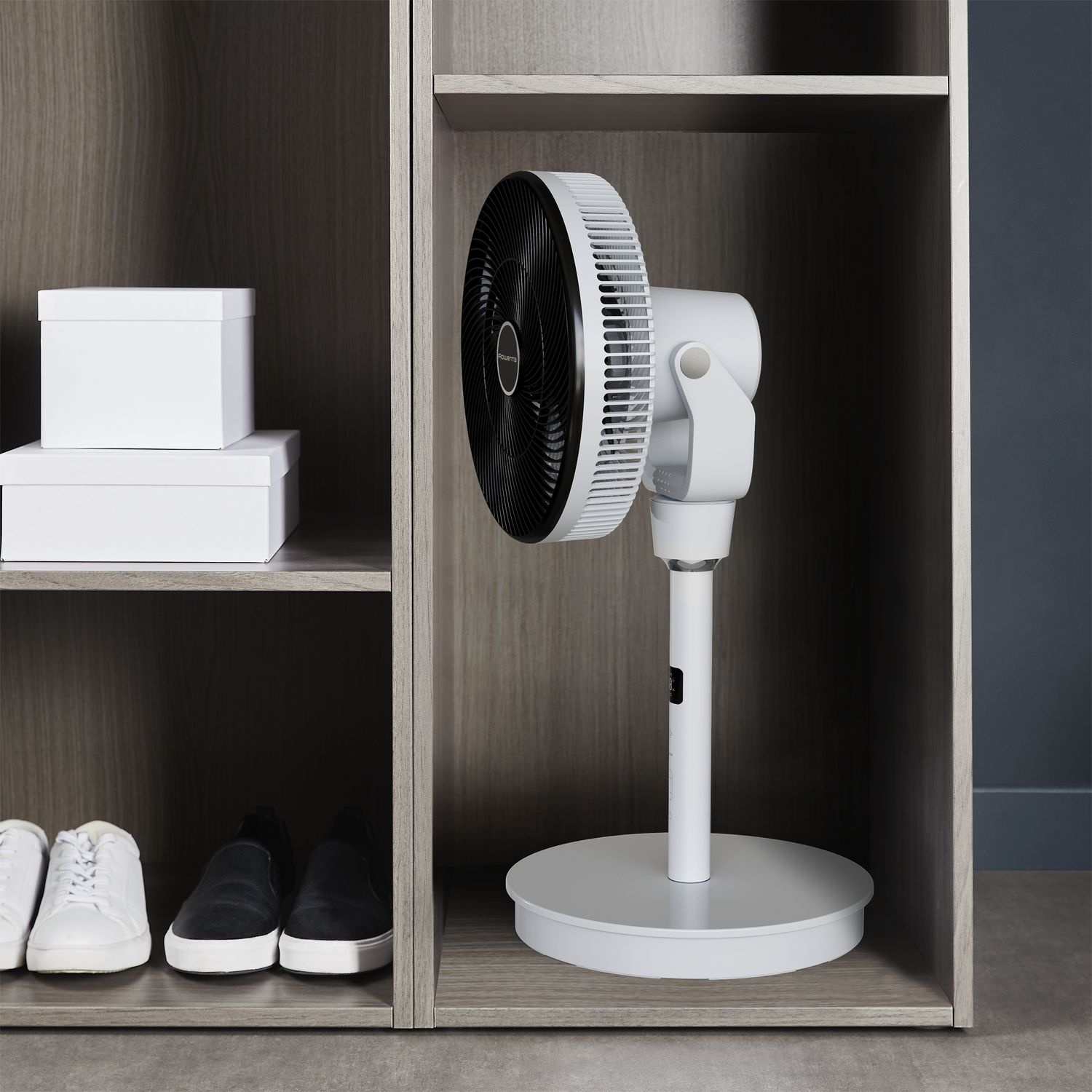 A white fan in a wooden cabinet. There are white boxes on the shelf above and shoes below.
